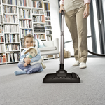 Person using a vacuum cleaner with a child sitting on the floor holding a teddy bear in a room with bookshelves.