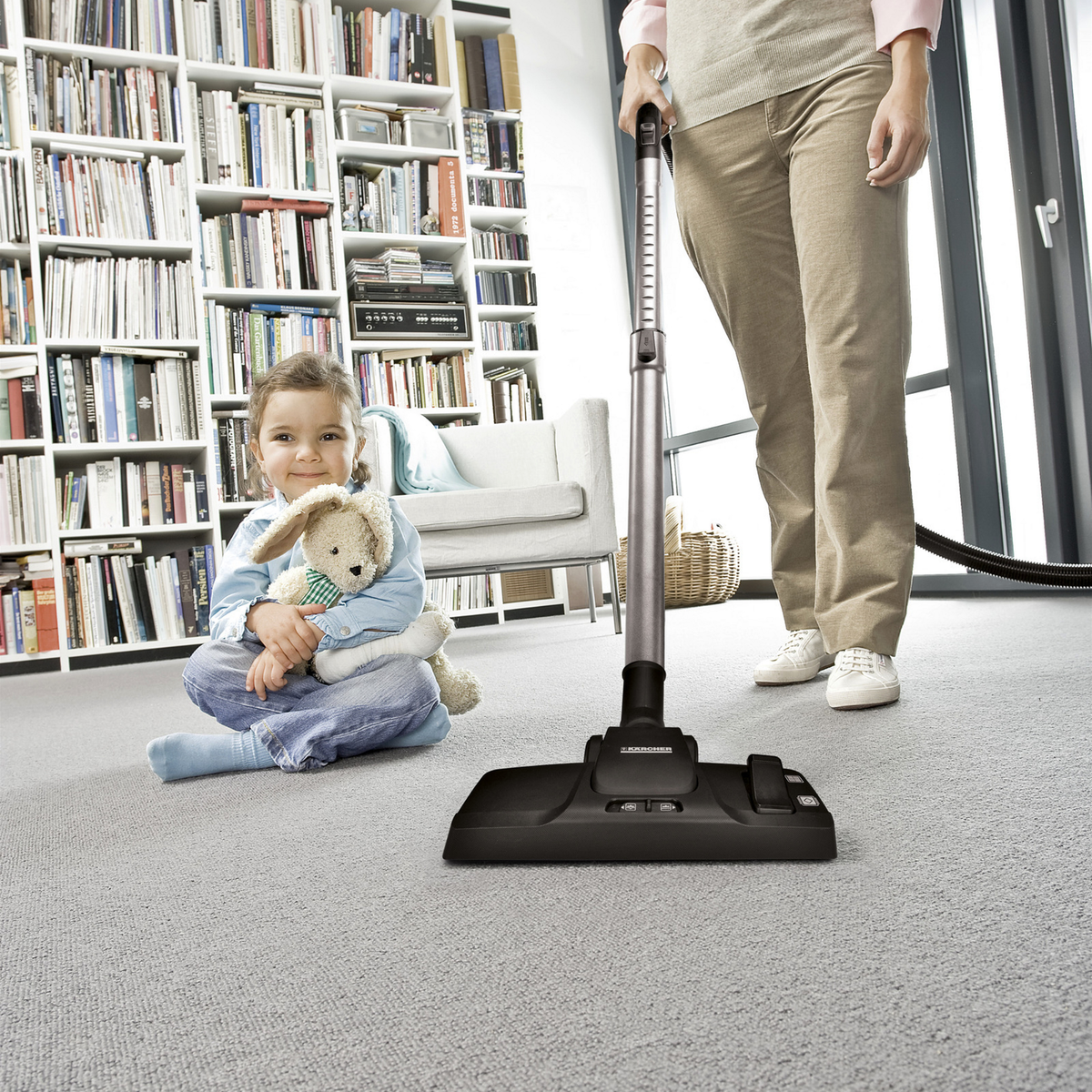 Person using a vacuum cleaner with a child sitting on the floor holding a teddy bear in a room with bookshelves.
