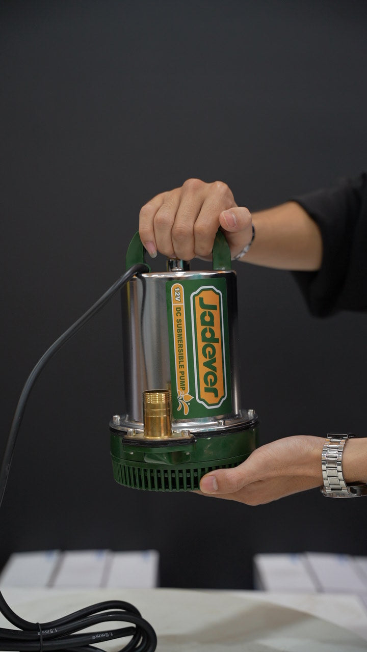 A person holds a green and silver submersible pump with a black cord against a dark gray background. The pump has a prominent green and yellow label with the brand name 'Hiebel'.