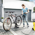 Woman washing a bicycle with a pressure washer in front of a modern house.