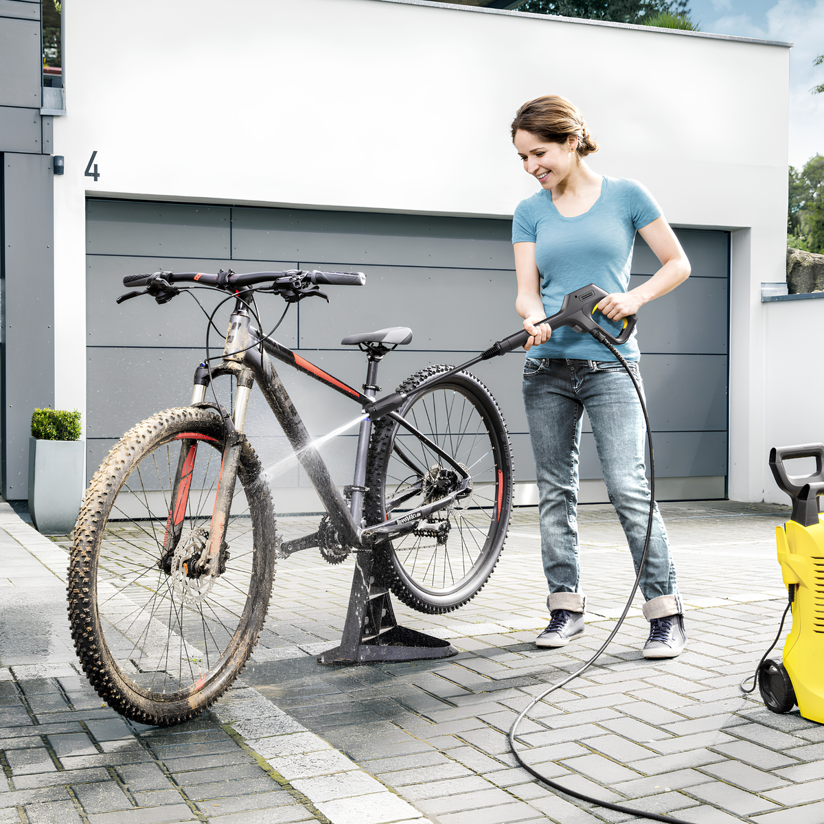 Woman washing a bicycle with a pressure washer in front of a modern house.