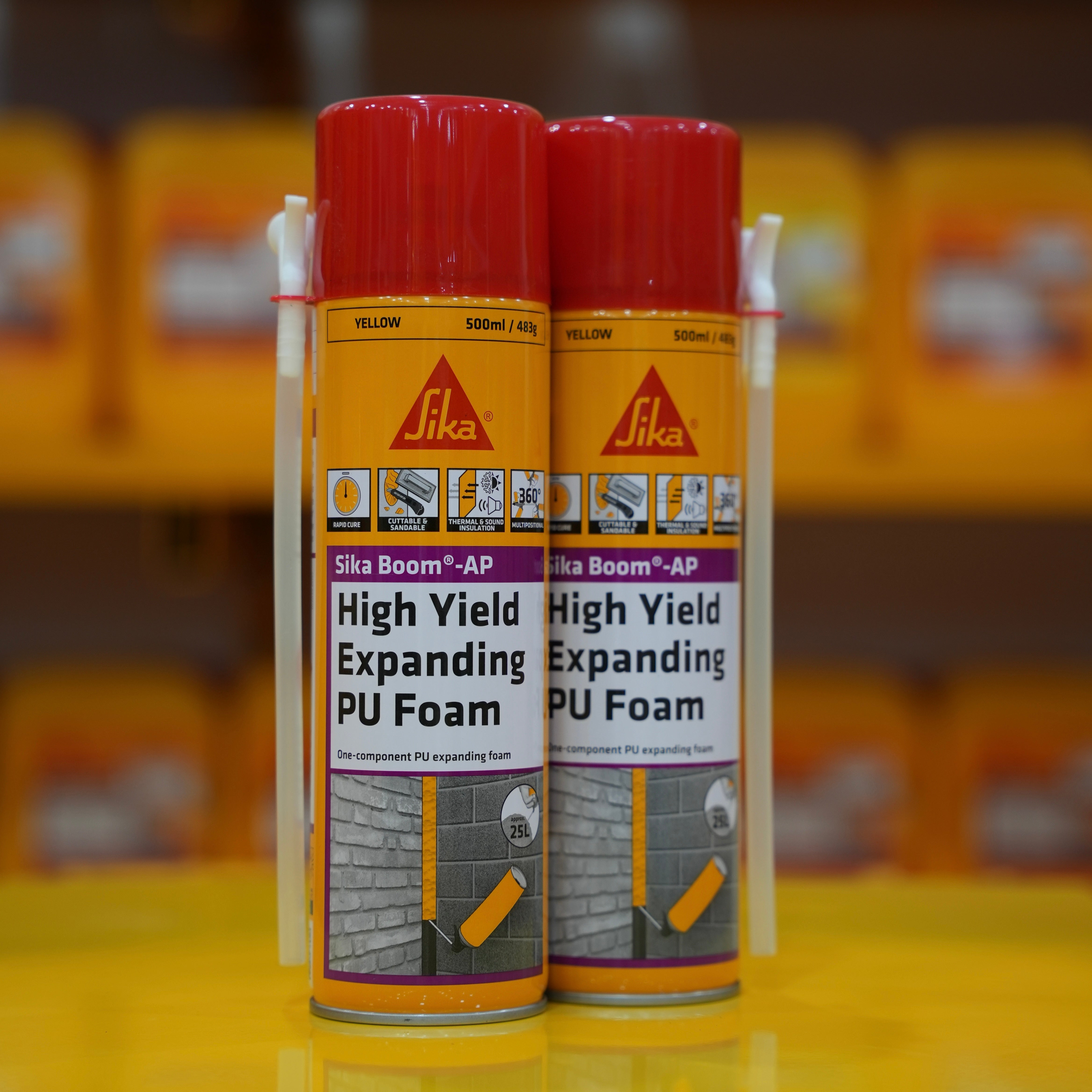 Two cans of Sika Boom AP High Yield Expanding PU Foam on a yellow surface with blurred background.